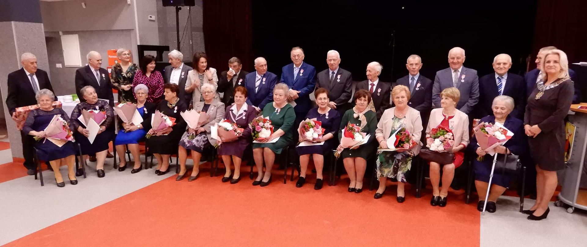 Na zdjęciu grupa ludzi. Panie siedzą na krzesłach i trzymają kwiaty. Mężczyźni stoją w drugim rzędzie. 