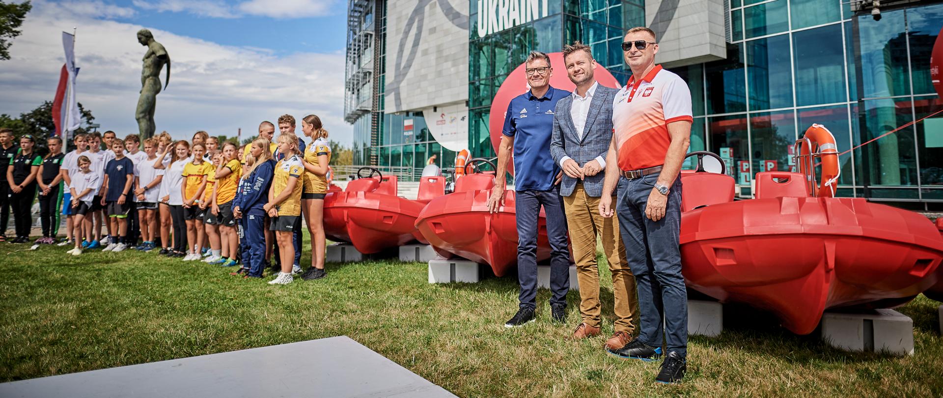 Minister Kamil Bortniczuk z prezesami Polskiego Związku Kajakowego i Polskiego Związku Towarzyszeń Wioślarskich stoi obok czerwonych motorówek zakupionych w ramach projektu Bezpieczna Woda. W tle dzieci trenujące sporty wodne