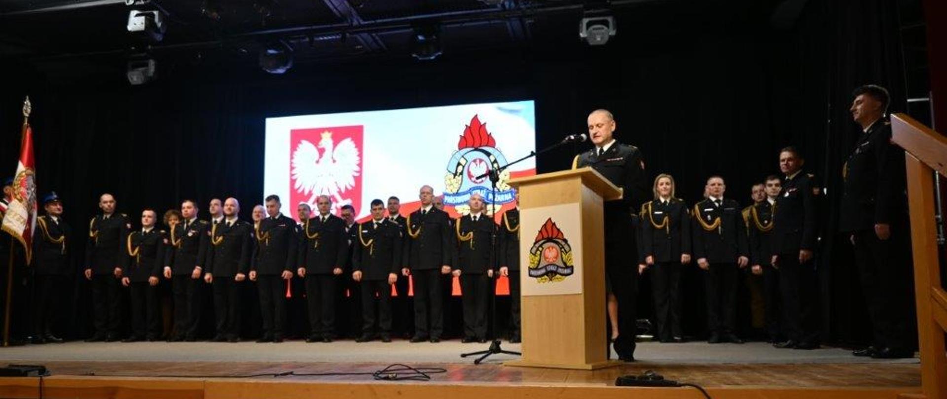 Na zdjęciu widać całą scenę z dużej odległości. Na środku sceny przy mównicy stoi strażak i przemawia. Za nim, przez całą szerokość sceny, stoi w długim rzędzie kilkunastu strażaków w czarnych mundurach galowych. Po lewej stronie sceny stoi poczet sztandarowy. Nad strażakami znajduje się wielki ekran z wyświetloną flagą Polski. Scena ma jasny, drewniany przód z pionowych listewek. U góry zdjęcia widać czarny sufit z zamontowanymi licznymi reflektorami. Po prawej stronie widać schody prowadzące na scenę.