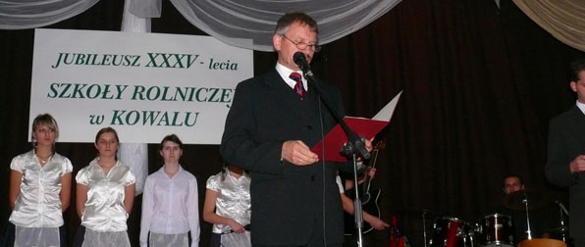 Na scenie przemawia dyrektor szkoły. w tle widać stojących uczniów oraz perkusjęzespołu muzycznego.