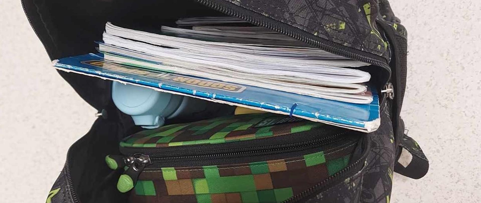 Na zdjęciu szkolny tornister. Tornister jest rozsunięty, tak że widzimy jego zawartość: duży piórnik zeszyty i książki oraz termos. Tornister koloru grafitowego w w geometryczne czarne i żółte wzory.