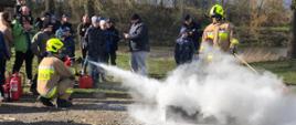 Na zdjęciu widać pokazy z gaszenia pożarów za pomocą gaśnicy. Dwóch strażaków udziela instruktażu praktycznego, a w tle widać osoby obserwujące proces gaszenia.