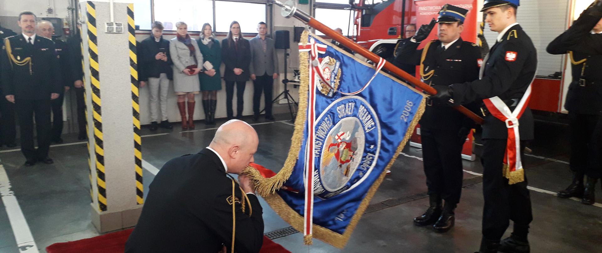 Komendant Powiatowy Państwowej Straży Pożarnej klęcząc na kolanie całuje sztandar Komendy Powiatowej Państwowej Straży Pożarnej w Dębicy. W tle zaproszeni goście i strażacy którzy są ubrani w mundury wyjściowe. Wszyscy znajdują się w pomieszczeniu garażowym straży pożarnej, przy czym w tym momencie znajduje się jeden samochód wewnątrz garażu.