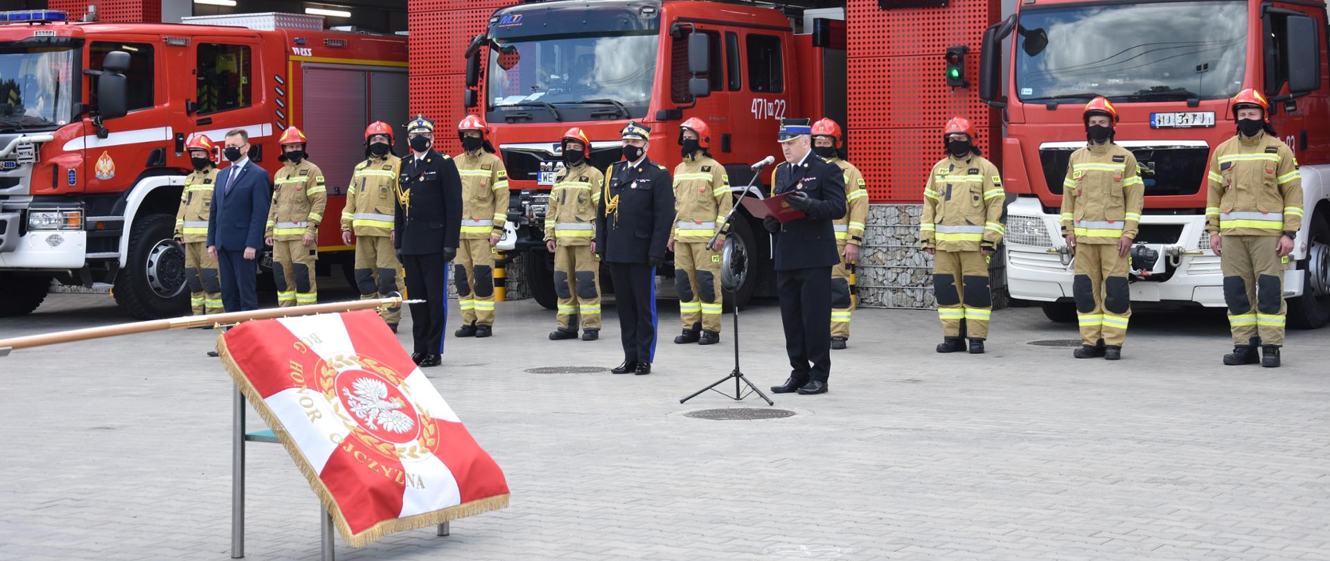 Zdjęcie z uroczystości, która odbyła się na dziedzińcu przed siedzibą komendy powiatowej PSP w Legionowie. Widoczne są wystawione trzy wozy strażackie, przed którymi stoją minister obrony narodowej, komendanci państwowej straży pożarnej oraz strażacy w żółtych mundurach. Na pierwszym planie zdjęcia widoczny sztandar.