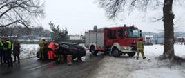 Samochód osobowy stoi w poprzek drogi. Wokół samochodu funkcjonariusze i ratownicy prowadzą działania ratownicze. W tle ustawiony w poprzek drogi samochód strażacki.