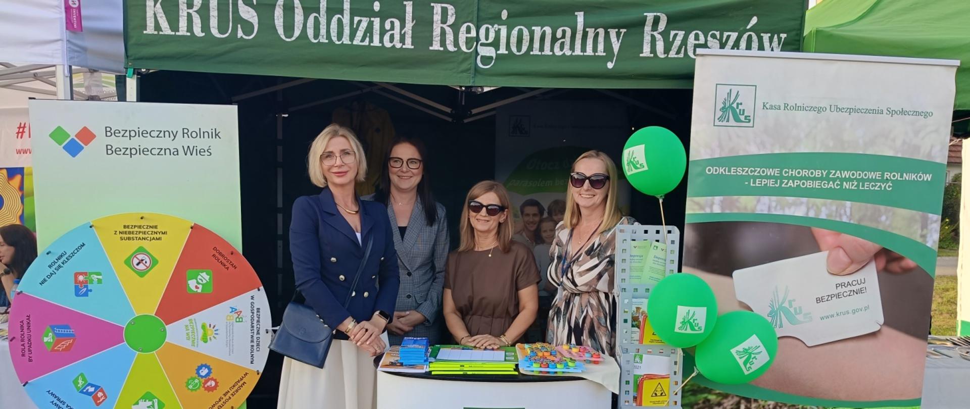 Przy stoisku informacyjno-prewencyjnym pod zielonym namiotem KRUS pozują do zdjęcia cztery uśmiechnięte kobiety. Po lewej stronie stoiska stoi plansza Koła Fortuny, po prawej – stojaki z materiałami informacyjnymi oraz plakat ostrzegający przed kleszczami. Stoisko udekorowane jest zielonymi balonami z logo KRUS.