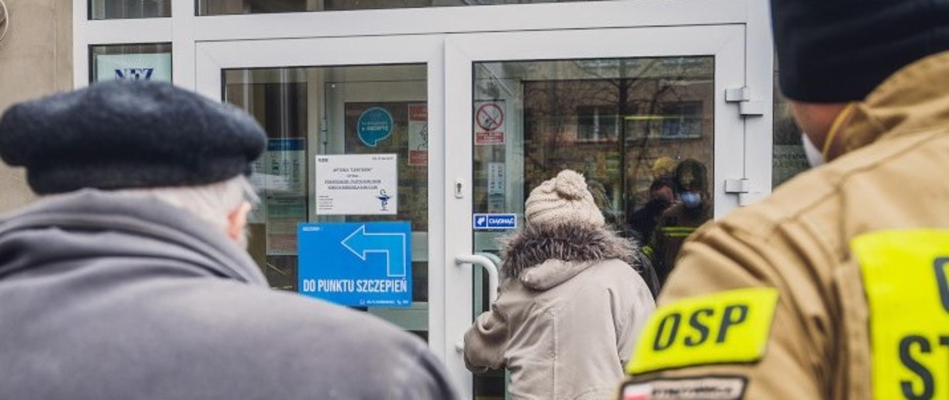 Baner - Dofinansowanie dla jednostek OSP ze środków Funduszu Przeciwdziałania COVID-19 " po prawej stronie w prawym rogu widać kawałek munduru strażaka z napisem OSP, na wprost kobieta wchodząca do punktu szczepień, po lewo starszy mężczyzna w czapce widok z tyłu