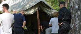 Zdjęcie przedstawia funkcjonariusza Komendy Powiatowej Państwowej Straży Pożarnej w Krośnie Odrzańskim, funkcjonariusza Komendy Powiatowej Policji w Krośnie Odrzańskim, Komendanta Obozu Harcerskiego „Struga” oraz harcerzy podczas sprawdzania procedur ewakuacji z terenu obozu. Zdjęcie wykonane zostało na tle namiotu w Obozie Harcerskim „Struga”.