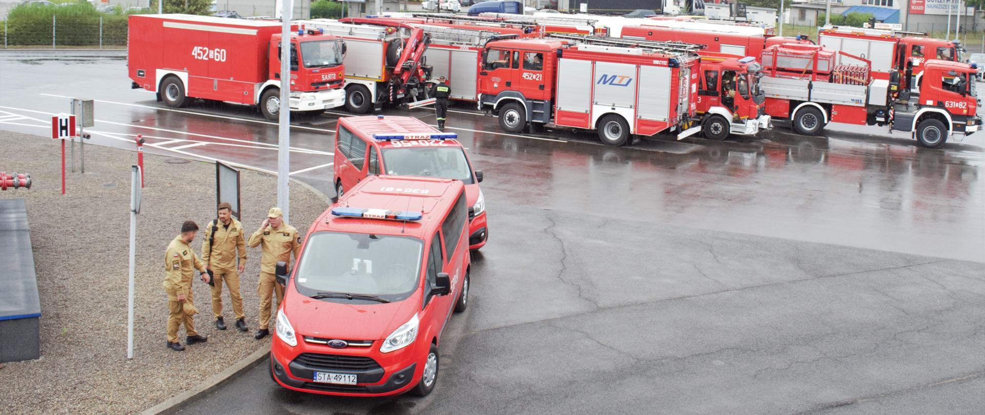 Na parkingu stoją czerwone samochody strażackie. Po lewej stronie zdjęcia obok czerwonego samochodu stoją trzej strażacy w mundurach w kolorze piaskowym.