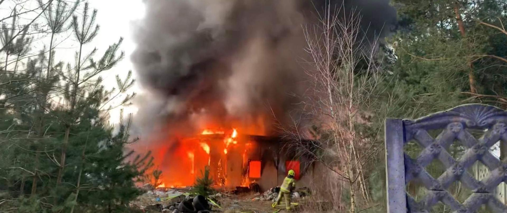 Dzień. Płonący żółto-czerwonym ogniem opuszczony budynek. Strażak przed nim rozwija linię gaśniczą. Duży czarny dym nad budynkiem.