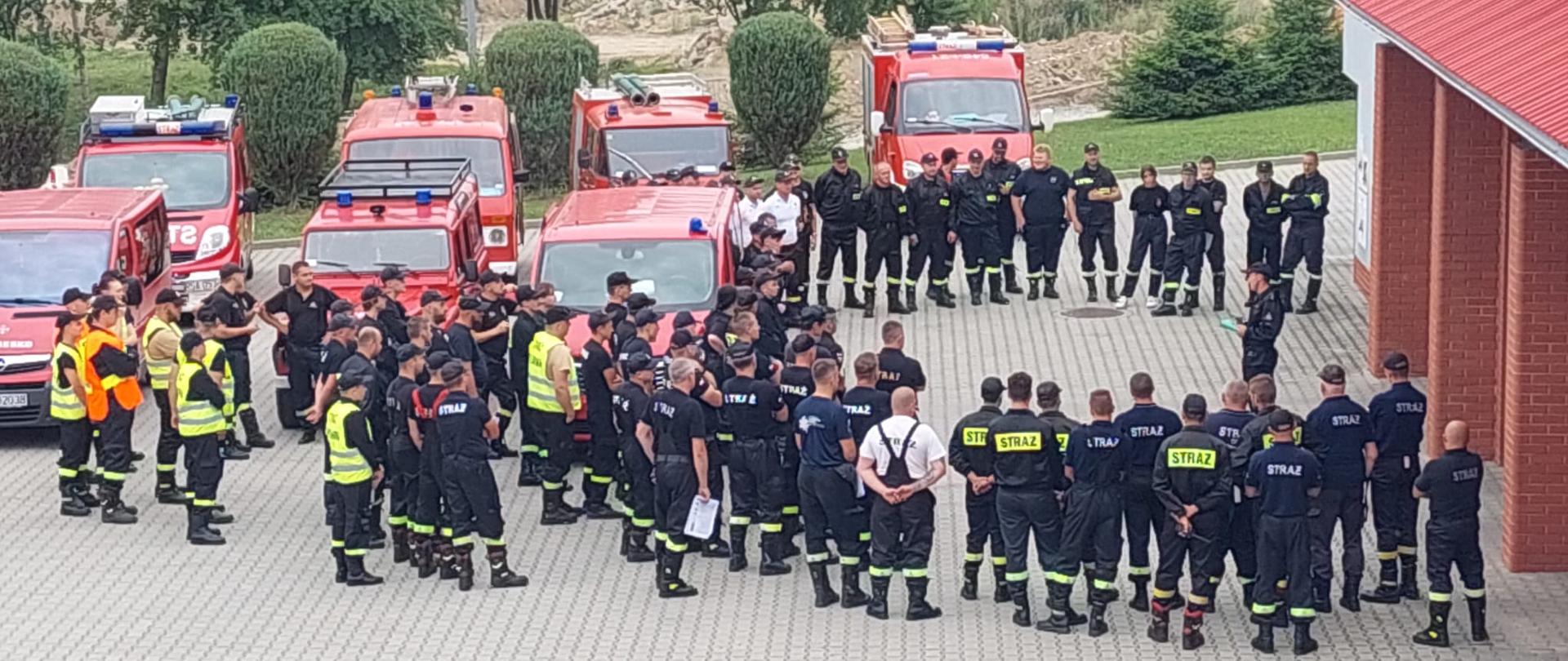 Zdjęcie przedstawia wiele samochodów strażackich i stojących na zbiórce strażaków (około 100 osób). Zdjęcie wykonane na zewnątrz na placu Komendy. 