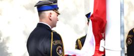 Strażacy w mundurach wyjściowych podpinają flagę państwową do masztu. Jeden oficer stoi bokiem do zdjęcia i podtrzymuje flagę państwową, strażak obok podnosi flagę na maszt. Widoczny emblemat na ramieniu strażaka z napisem Kompania Reprezentacyjna Państwowej Straży Pożarnej.