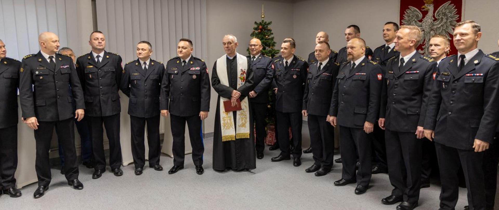Strażacy Państwowej Straży Pożarnej w mundurach wyjściowych w tym nadbryg. Krzysztof Kędryk Opolski Komendant Wojewódzki Państwowej Straży Pożarnej oraz ksiądz Tomasz Józkowicz stoją na świetlicy Ośrodka Szkolenia Komendy Wojewódzkiej Państwowej Straży Pożarnej w Opolu. W tle widoczna ustrojona choinka oraz Godło Rzeczypospolitej Polskiej.