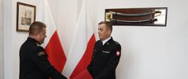 Funkcjonariusz Państwowej Straży Pożarnej wręcza czerwoną teczkę strażakowi mężczyźni ubrani są mundury wyjściowe za nimi stoją dwie flagi Polski na ścianach wiszę obraz oraz szabla. 