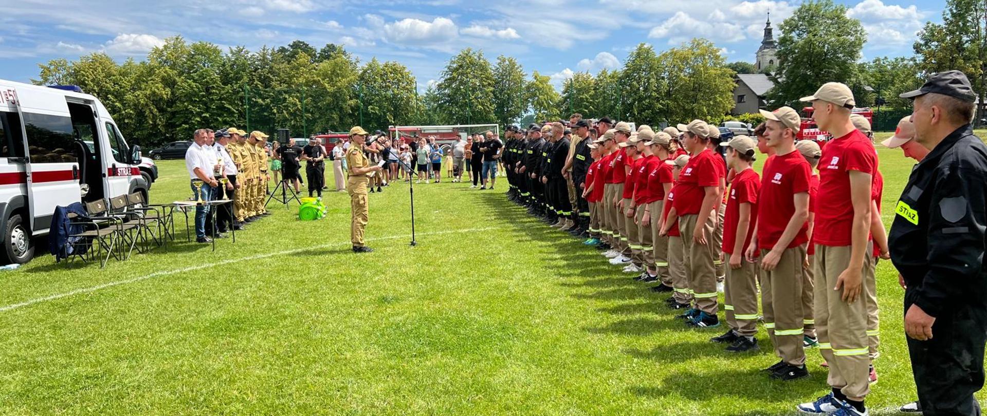 Zdjęcie przedstawia strażaków OSP oraz drużynę MDP ustawionych po prawej stronie w dwuszeregu. Ubrani są w ubranie koszarowe, pasy strażackie oraz hełmy. Strażacy stoją na boisku z trawą. Po lewej znajduje się samochód pożarniczy oraz kadra sędziowska i wójtowie gmin.