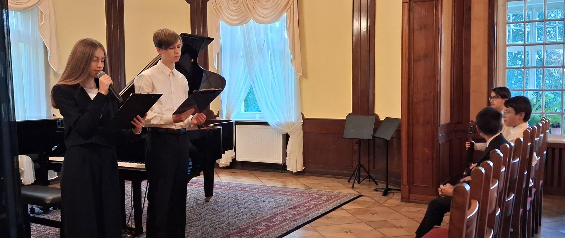 Zdjęcie kolorowe. Elegancka sala koncertowa wykończona drewnianymi boazeriami. Po lewej stronie, obok czarnego fortepianu , stoi dwoje młodych ludzi prowadzących wydarzenie. Dziewczyna po lewej, ubrana w czarny garnitur, trzyma czarną podkładkę z kartkami i przemawia do mikrofonu. Chłopak po prawej, w białej koszuli, czarnych spodniach i pod krawatem, trzyma podobną podkładkę i patrzy na tekst. Po prawej stronie, na drewnianych krzesłach z czerwonymi tapicerowanymi siedziskami, siedzą słuchacze. W tle widać jasne ściany, okna z białymi firankami oraz ozdobny żyrandol.