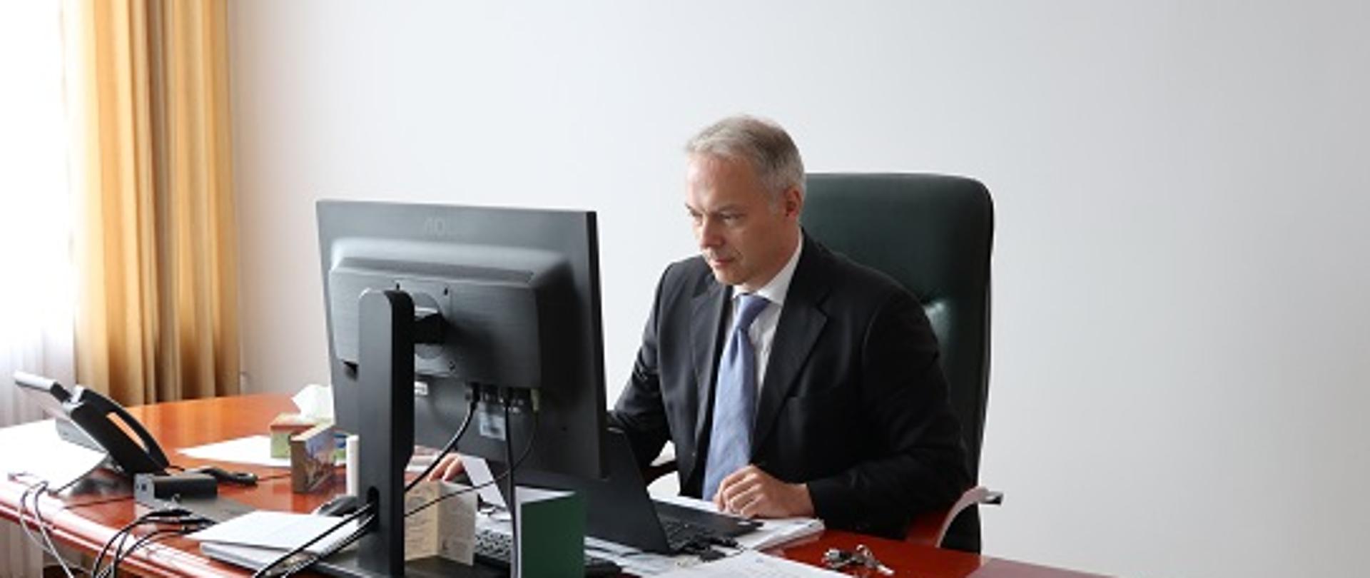 Wiceminister Jacek Żalek siedzi przy biurku, przed komputerem. Na biurku widoczne dokumenty.