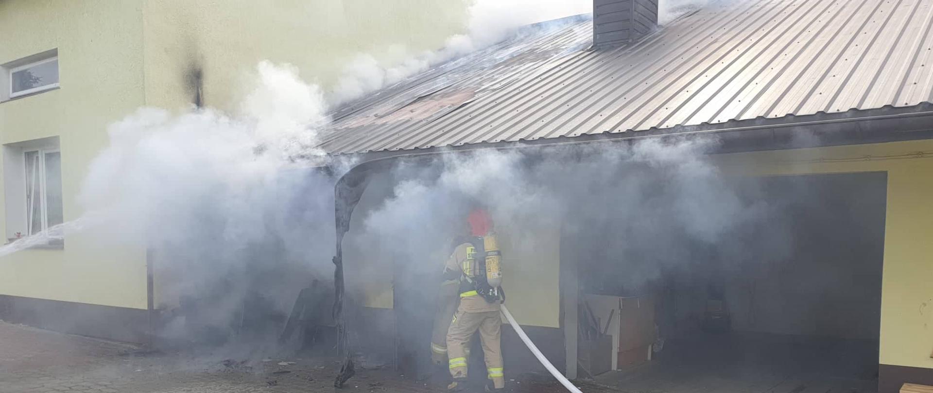 Na zdjęciu strażaków ubranych w sprzęt ochrony układu oddechowego podających prąd wody na palące się poddasze budynku gospodarczego . Do budynku gospodarczego przylega budynek mieszkalny. Panuje silne zadymienie na poddaszu budynku gospodarczego.