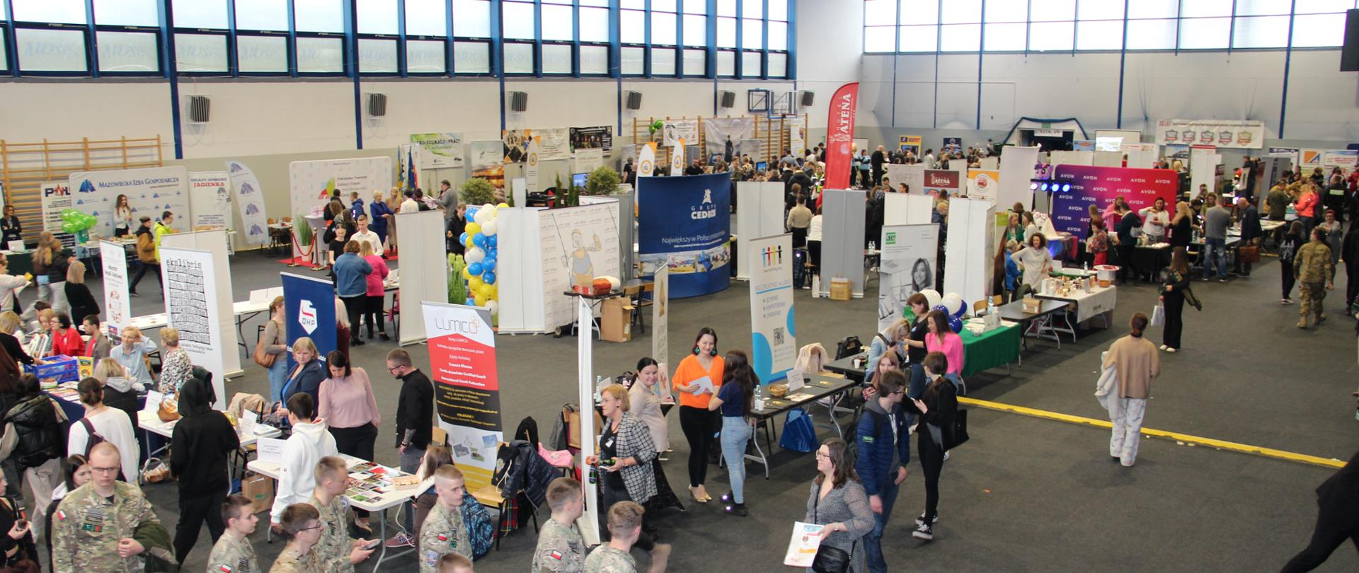 Na zdjęciu grupa ludzi spaceruje po dużej hali z różnymi stanowiskami 