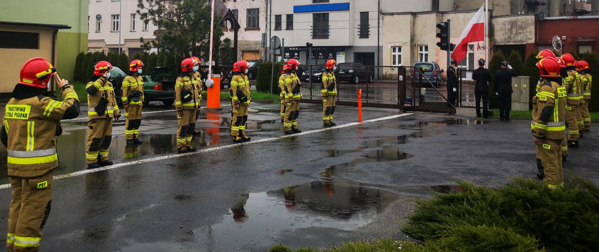 Zdjęcie wykonano podczas uroczystego podniesienia flagi państwowej na maszt na palcu Komendy Powiatowej PSP w Dniu Flagi Rzeczypospolitej Polskiej. Uroczystego podniesienia dokonuje poczet sztandarowy w obecności strażaków ubranych w ubrania specjalne.