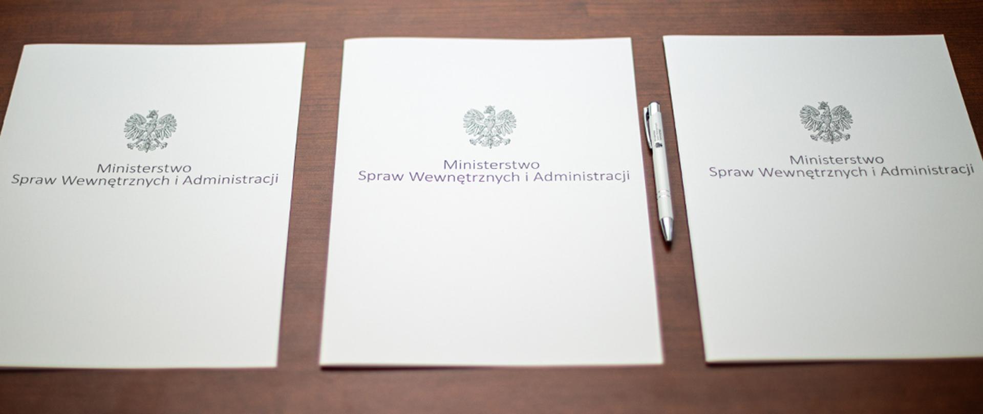 Na zdjęciu widać trzy leżące w rzędzie teczki na dokumenty z napisem "Ministerstwo Spraw Wewnętrznych i Administracji" i orłem z godła narodowego. Obok jednej z nich leży długopis MSWiA.