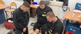 Na zdjęciu trzech druhów z Ochotniczych Straży Pożarnych z powiatu gorlickiego ubrani w czarne ubrania koszarowe w sali konferencyjnej w Komendzie Powiatowej Państwowej Straży Pożarnej podczas recertyfikacji uprawnień z zakresu kwalifikowanej pierwszej pomocy wykonujący czynności ratownicze z użyciem fantomu.