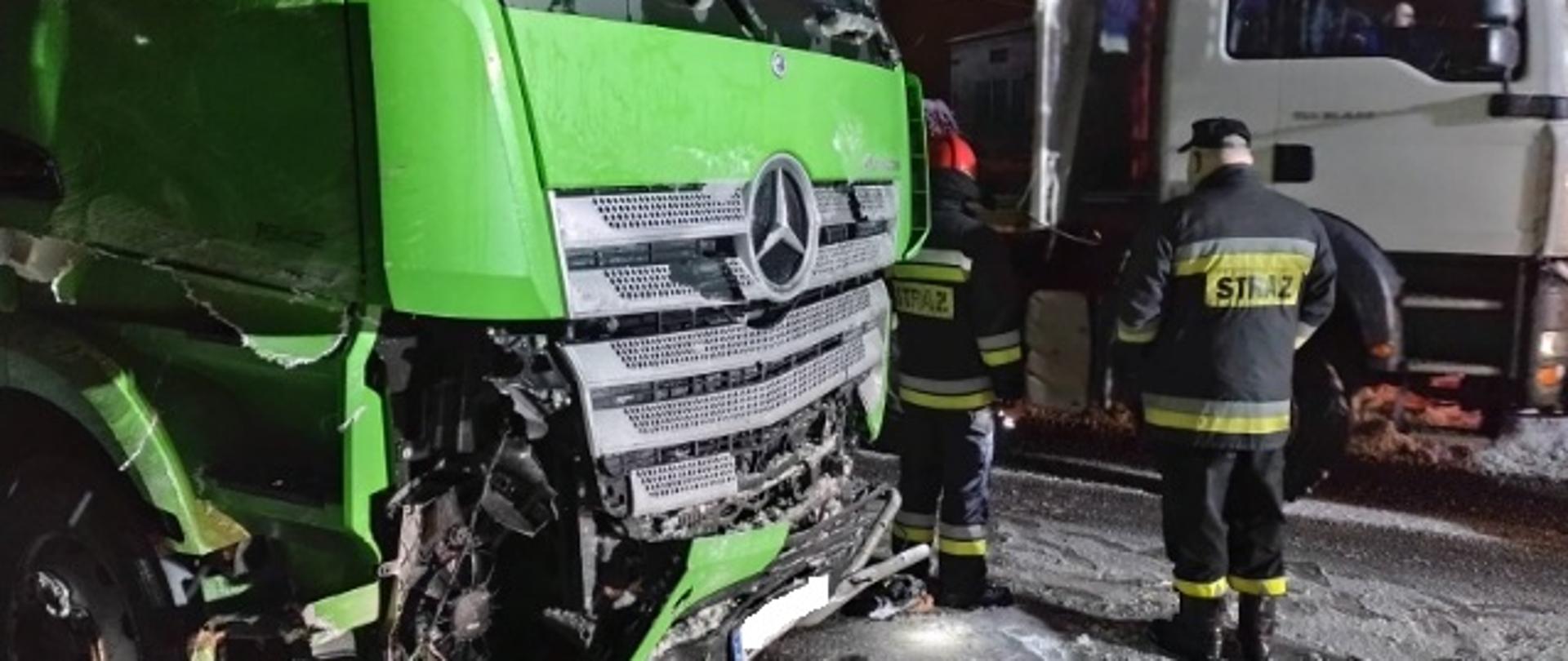 Na pierwszym planie widzimy przód uszkodzonego samochodu ciężarowego, w tle dwóch strażaków. 