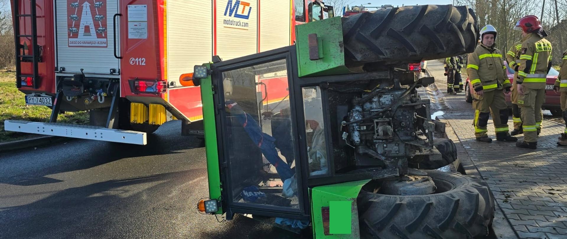 Ciągnik rolniczy leży na boku na drodze, po prawej samochód strażacki