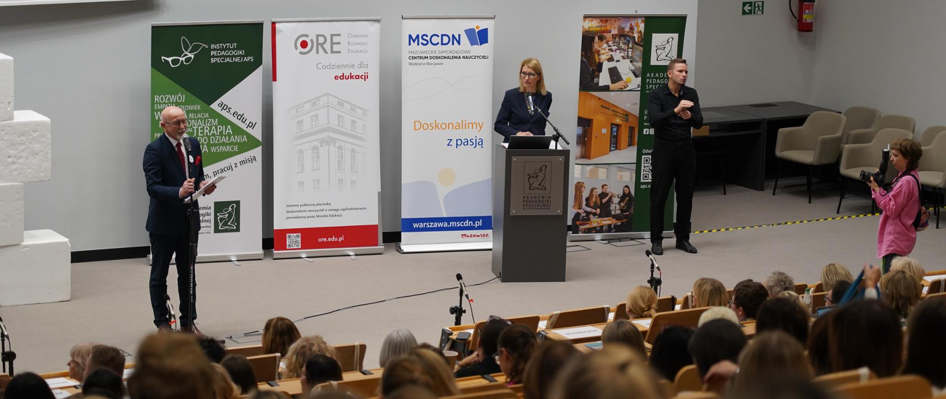 Zdjęcie z sali audytoryjnej podczas konferencji. Na pierwszym planie widoczna jest duża grupa uczestników siedzących tyłem do kamery. Na scenie znajdują się trzy roll-upy z logo organizatorów: Instytutu Pedagogiki Specjalnej APS, ORE i MSCDN. Po lewej stronie mównicy stoi mężczyzna trzymający dokumenty, a po prawej stoi kobieta przemawiająca z mównicy. Obok niej, na prawo, stoi tłumacz języka migowego.