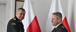 Pomorski komendant wojewódzki Państwowej Straży Pożarnej ściska dłoń strażakowi trzymającemu czerwoną teczkę. Funkcjonariusze Państwowej Straży Pożarnej ubrani w mundury wyjściowe z białą koszulą. Za nimi ustawione są dwie flagi Polski na ścianie zawieszony jest obraz. 
