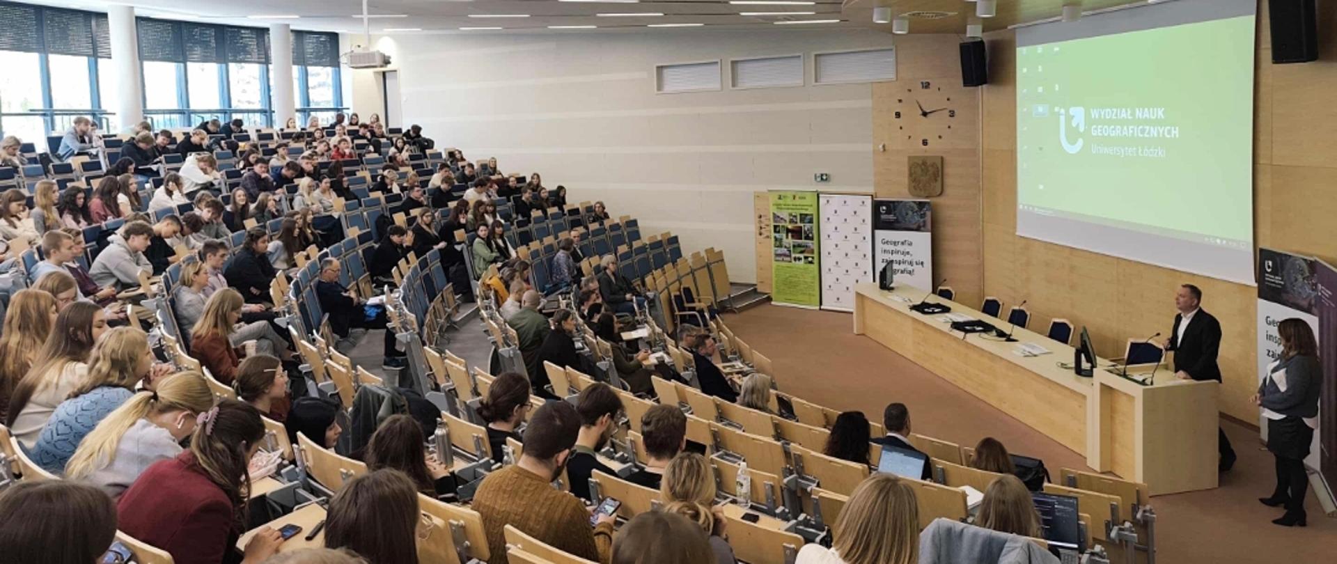 aula, grupa ludzi widoczna z boku, na dole meżczyzna w garniturze przemawia do zebranych, zanim na dużym ekranie napis na zielonym tle: Wydział Nauk Geograficznych Uniwersytet Łódzki 