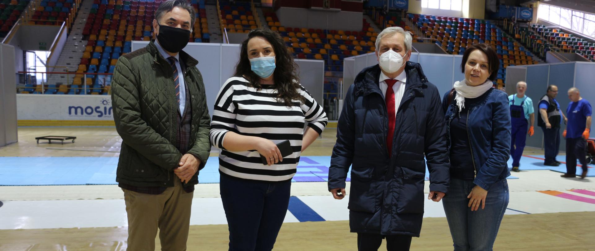 Na zdjęciu cztery osoby, stojące na tafli nieczynnego lodowiska. Od lewej dyrektor opolskiego NFZ, pracownica Toropolu, wojewoda opolski, oraz pracownica Toropolu. W tle postęp orac nad przygotowywaniem punkty tj. plastikowe boksy, gdzie będą szczepieni opolanie przeciwko koronawirusowi. 
