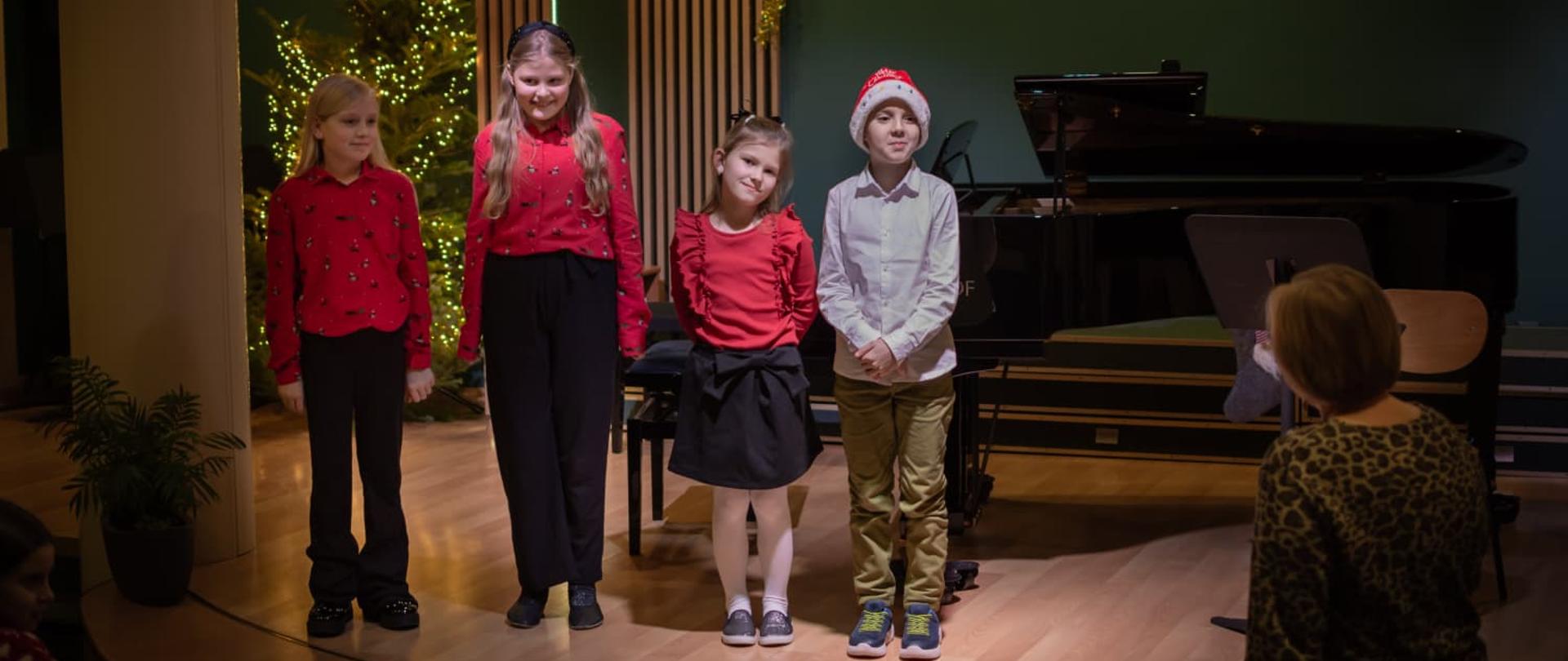 Na scenie widać uczniów którzy są ubrani piękne czerwone bluzki oraz w białą koszulę i uśmiechają się do publiczności w tle widoczny jest fortepian oraz ozdoby świąteczny