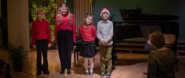 Na scenie widać uczniów którzy są ubrani piękne czerwone bluzki oraz w białą koszulę i uśmiechają się do publiczności w tle widoczny jest fortepian oraz ozdoby świąteczny