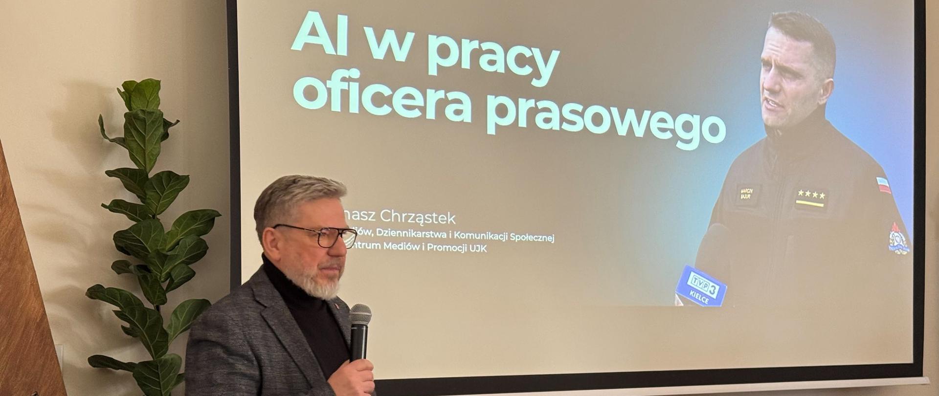 Mężczyzna w okularach, ubrany w ciemną marynarkę i czarny golf, stoi przed ekranem projekcyjnym i trzyma mikrofon, prowadząc prezentację. Na ekranie widoczny jest slajd z tytułem „AI w pracy oficera prasowego” oraz imieniem i nazwiskiem prelegenta, a także zdjęcie strażaka udzielającego wypowiedzi mediom. Przed ekranem znajduje się laptop podłączony do sprzętu multimedialnego. Wnętrze ma charakter konferencyjny, z klasycznymi meblami i rośliną dekoracyjną po lewej stronie. Zdjęcie przedstawia szkolenie lub wykład dotyczący wykorzystania sztucznej inteligencji w pracy komunikacyjnej.