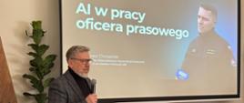 Mężczyzna w okularach, ubrany w ciemną marynarkę i czarny golf, stoi przed ekranem projekcyjnym i trzyma mikrofon, prowadząc prezentację. Na ekranie widoczny jest slajd z tytułem „AI w pracy oficera prasowego” oraz imieniem i nazwiskiem prelegenta, a także zdjęcie strażaka udzielającego wypowiedzi mediom. Przed ekranem znajduje się laptop podłączony do sprzętu multimedialnego. Wnętrze ma charakter konferencyjny, z klasycznymi meblami i rośliną dekoracyjną po lewej stronie. Zdjęcie przedstawia szkolenie lub wykład dotyczący wykorzystania sztucznej inteligencji w pracy komunikacyjnej.