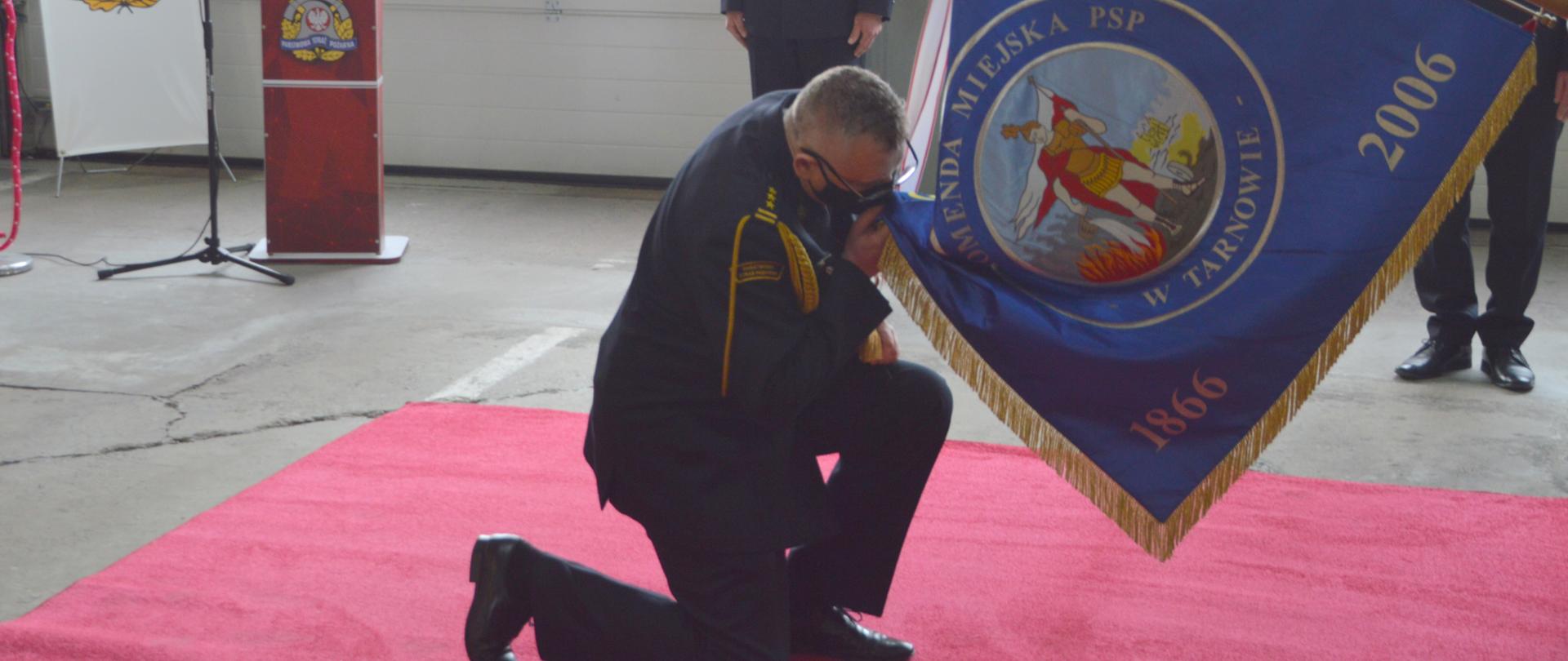 Zdjęcie przedstawia Komendanta Miejskiego PSP w Tarnowie w chwili pożegnania ze sztandarem KM PSP w Tarnowie. Komendant całuje sztandar przyklękając na prawe kolano w tle widoczni są oficerowie KM PSP Tarnów.