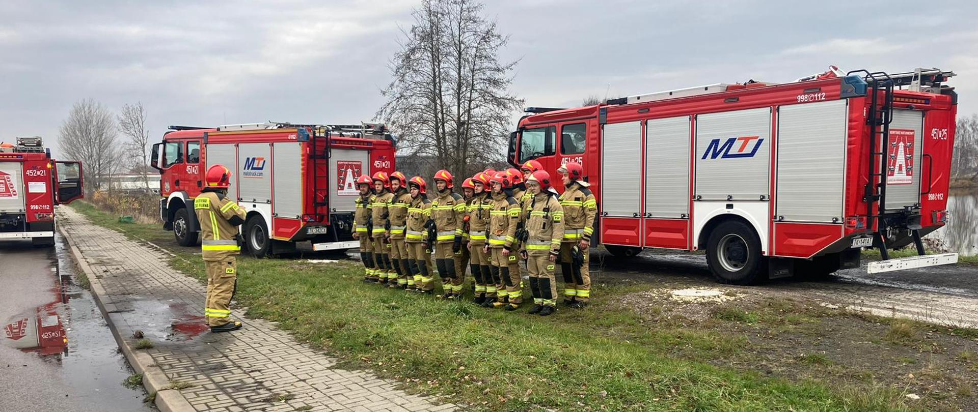 Na zdjęciu widoczni są strażacy w trakcie zbiórki na której Dowódca omawia założenie ćwiczeń oraz cele do osiągnięcia. W tle widoczne samochody ratowniczo gaśnicze.