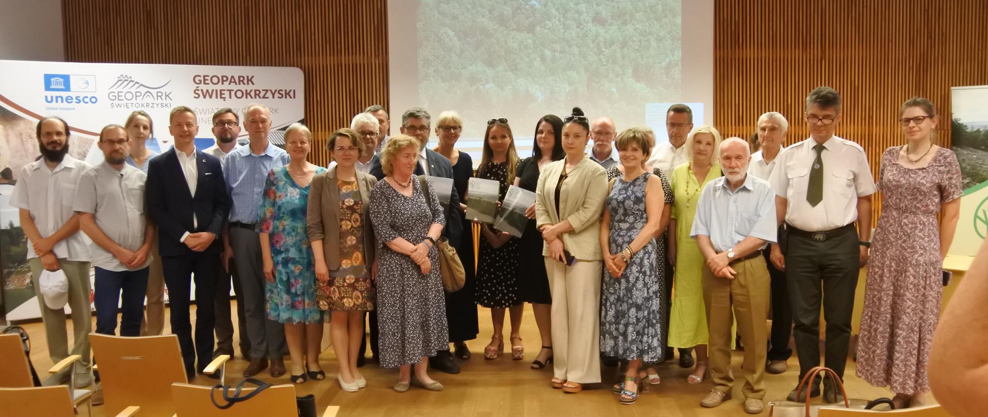 Zdjęcie przedstawia dużą grupę osób pozujących do wspólnej fotografii w nowoczesnej sali konferencyjnej lub auli. Uczestnicy spotkania stoją w rzędzie, są ubrani w stroje wizytowe oraz letnie i uśmiechają się w stronę obiektywu. W tle widoczna jest drewniana ściana, duży ekran projekcyjny z wyświetlonym obrazem oraz baner reklamowy z logo UNESCO Geopark Świętokrzyski. Na pierwszym planie po lewej i prawej stronie widać puste krzesła konferencyjne ustawione na jasnej, drewnianej podłodze.