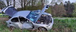 Samochód osobowy znajduje się na poboczu drogi. Karoseria uszkodzona w wyniku wypadku. Za pojazdem na jezdni stoi radiowóz policji. W tle las.