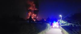Zdjęcie przedstawia wóz strażacki jadący do pożaru. W tle widać czerwoną łunę oraz czarny dym.