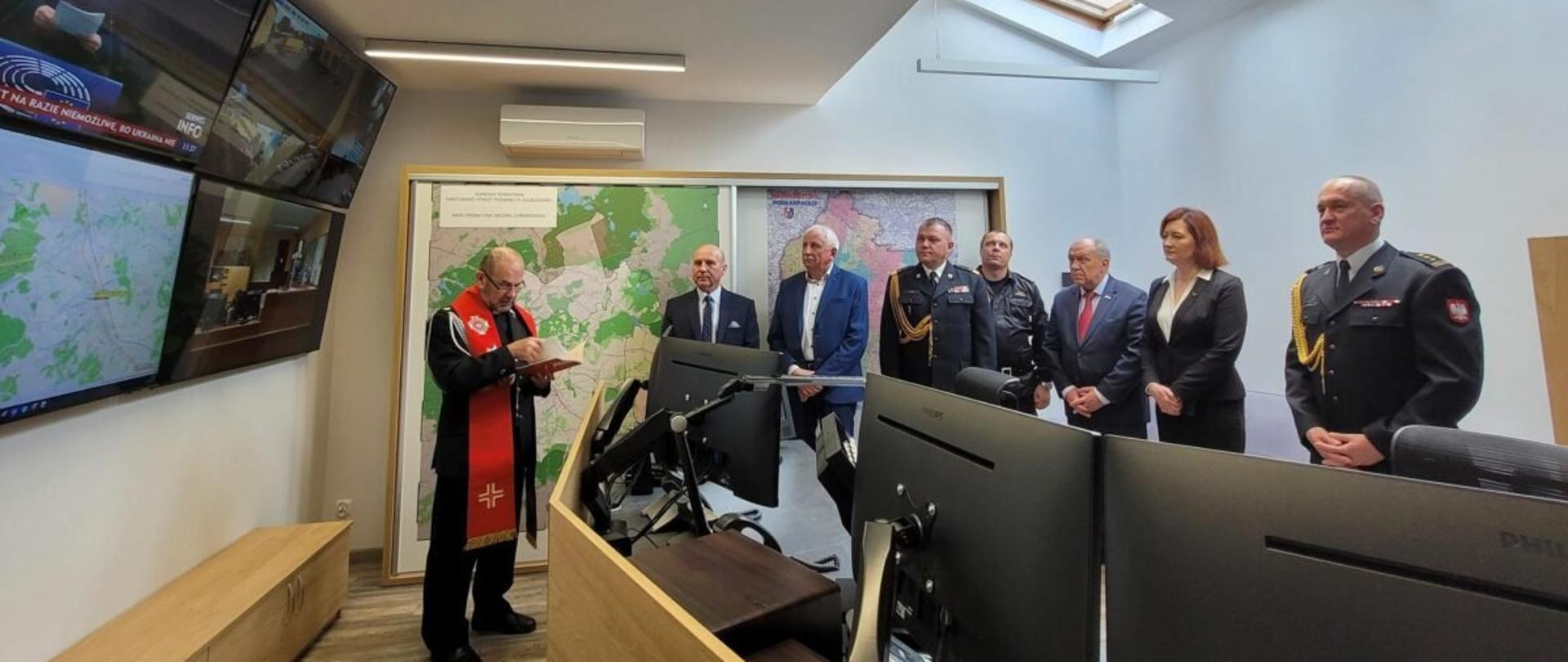 Zdjęcie wykonane wewnątrz pomieszczenia. W pomieszczeniu po lewej stronie umieszczone na ścianie są ekrany telewizyjne i komputerowe. W pomieszczeniu stoi grupa osób biorących udział w uroczystości. Wśród nich oficerowie Państwowej Straży Pożarnej w mundurach galowych.