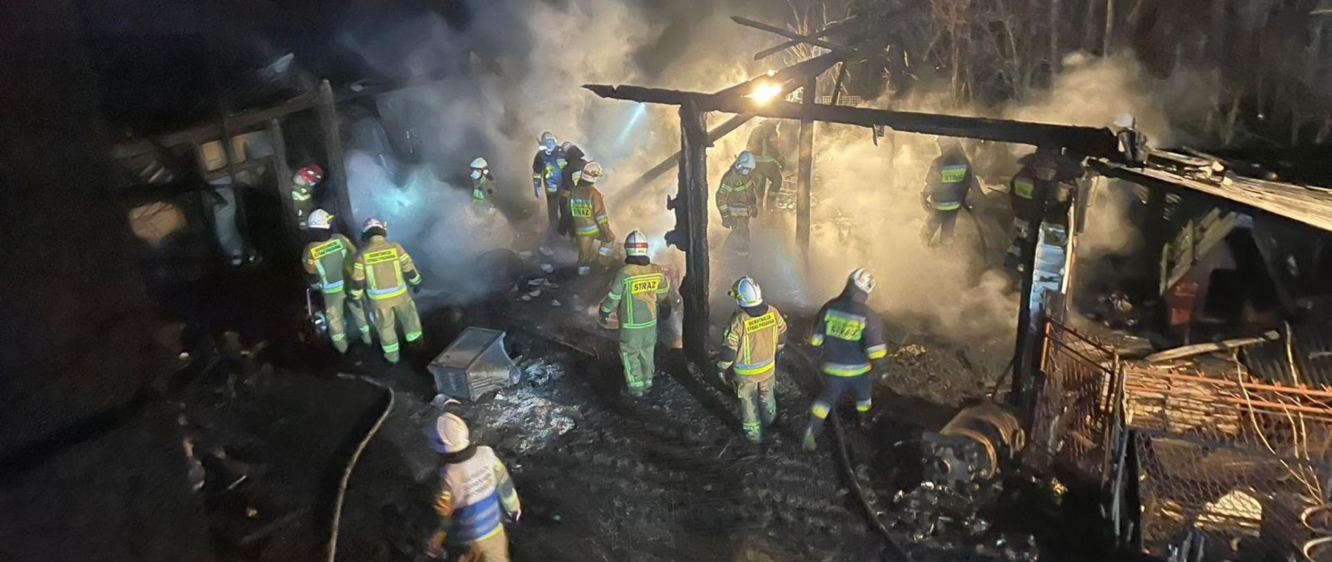 Noc. Widok z góry na podwórze. Po prawej widoczna spalona drewniana konstrukcja budynku gospodarczego. Po lewej w głębi widoczna konstrukcja drugiego budynku. Na podwórzu kilkunastu strażaków w ubraniach specjalnych i hełmach. Na ziemi widoczne węże strażackie.