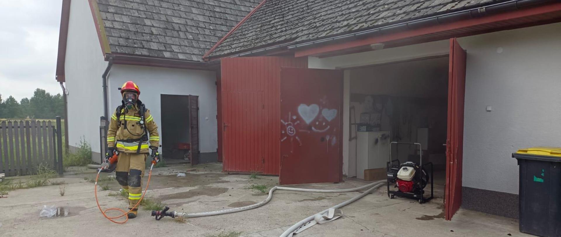 Na zdjęciu widać budynek gospodarczy oraz rozwiniete przed budynkiem linie gaśnicze, strażak z narzędziami hydraulicznymi, w drzwiach budynku wstawiony wentylator, trwa oddymianie pomieszczeń.
