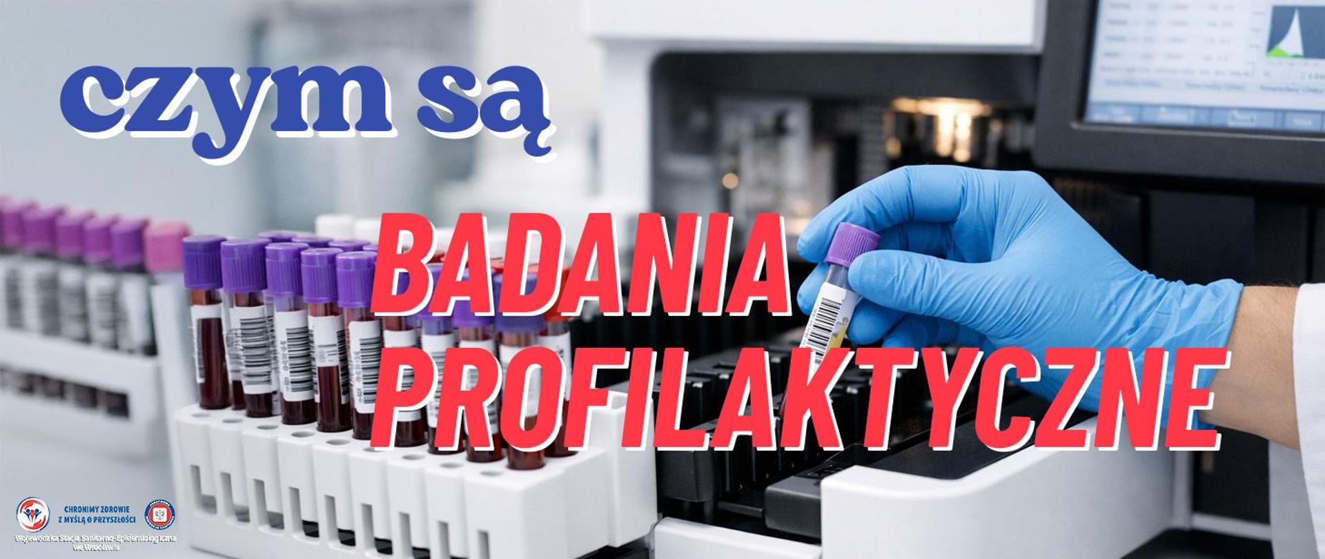 Technik laboratoryjny w niebieskich rękawiczkach trzyma probówkę z krwią i wkłada ją do automatycznego analizatora, w tle stoją probówki w stojaku, a ekran analizatora pokazuje wykresy wyników.