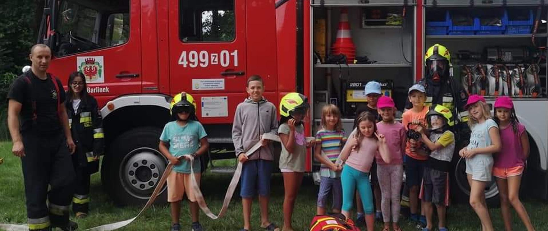 Na zdjęciu grupa dzieci przymierzających ubrania strażaka. Obok strażak i pojazd gaśniczy.