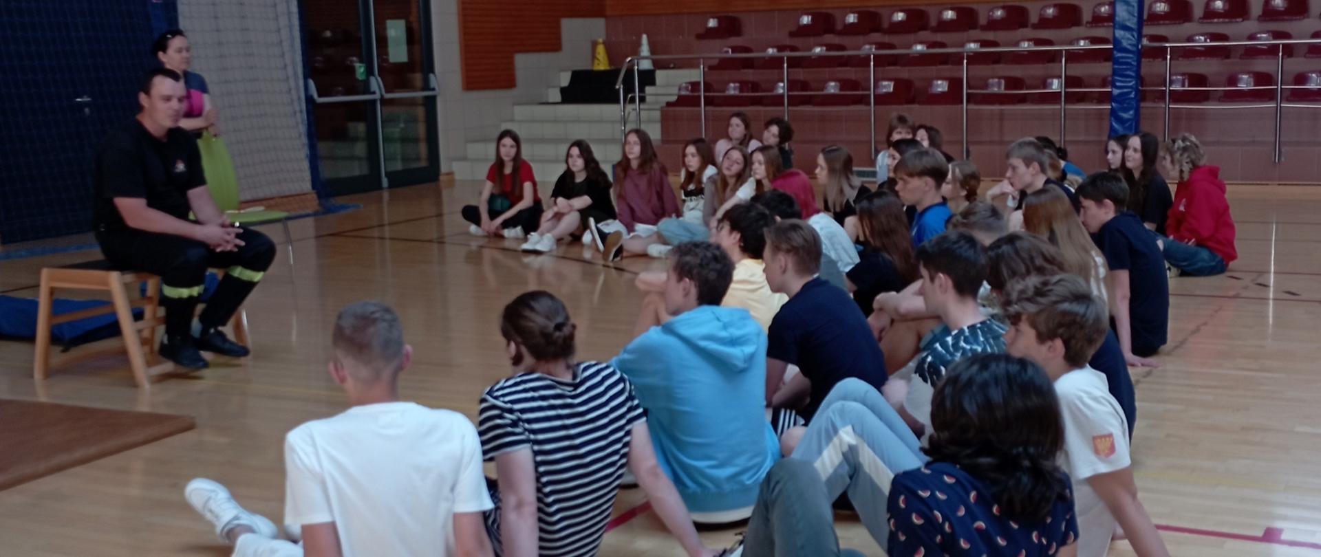 Na zdjęciu przedstawiono warsztaty z uczniami w zespole szkół w ramach profilaktyki bezpieczeństwa. Ratownik na sali gimnastycznej prowadzi prelekcję, a młodzież szkolna siedzi na parkiecie słuchając.