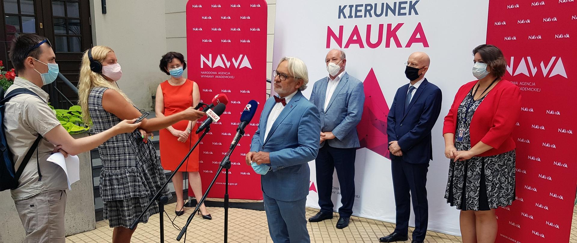 Wiceminister Maksymowicz przemawia do mikrofonu, za nim ściana z kogiem NAWA i napisem Kierunek Nauka.