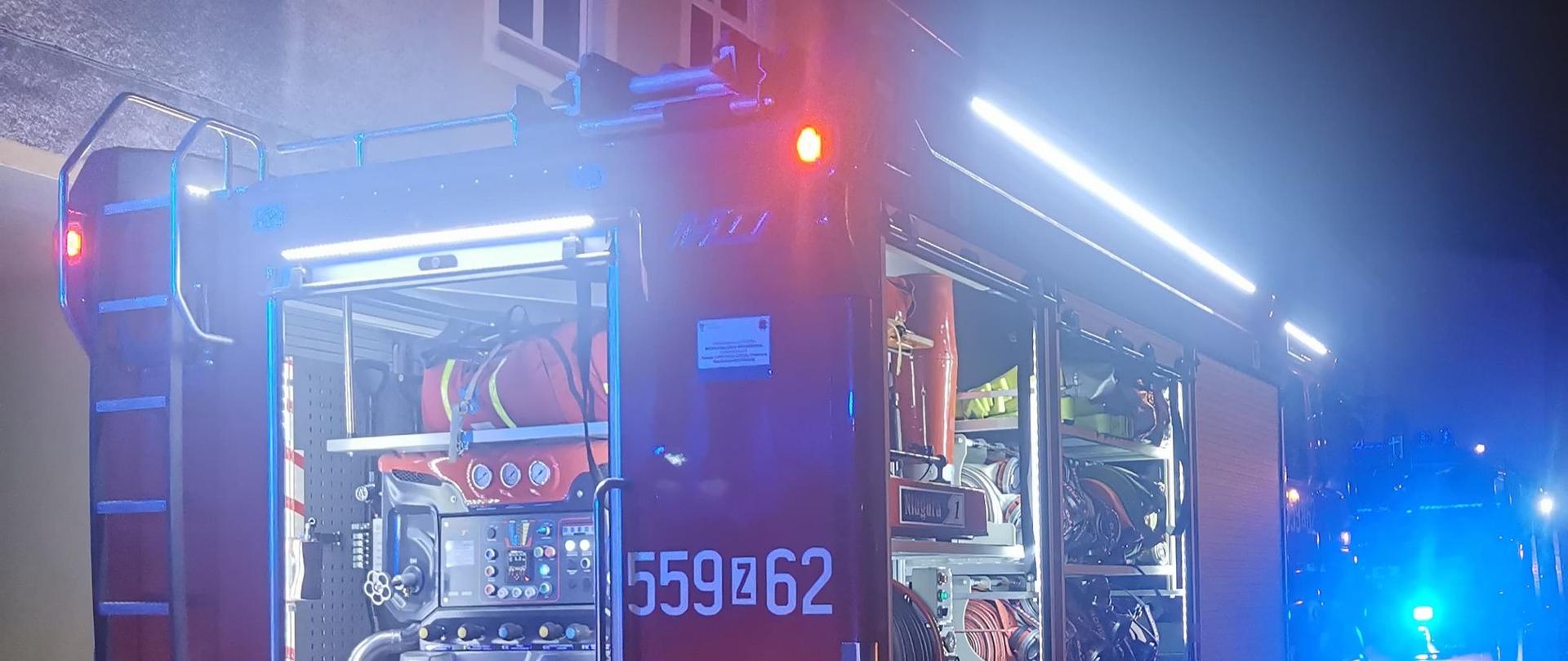 Zdjęcie przedstawia tył średniego samochodu ratowniczo-gaśniczy straży pożarnej z otwartymi oświetlonymi skrytkami, włączonymi światłami pozycyjnymi oraz niebieskimi błyskowymi. W tylnej skrytce widać autopompę samochodu strażackiego, który podaję wodę do pożaru. Z lewej strony widać czerwony wąż podłączony do nasady tłocznej pojazdu. Z prawej strony uchwycono część samochodu osobowego, zaś z tyłu widać kolejny wóz strażacki.