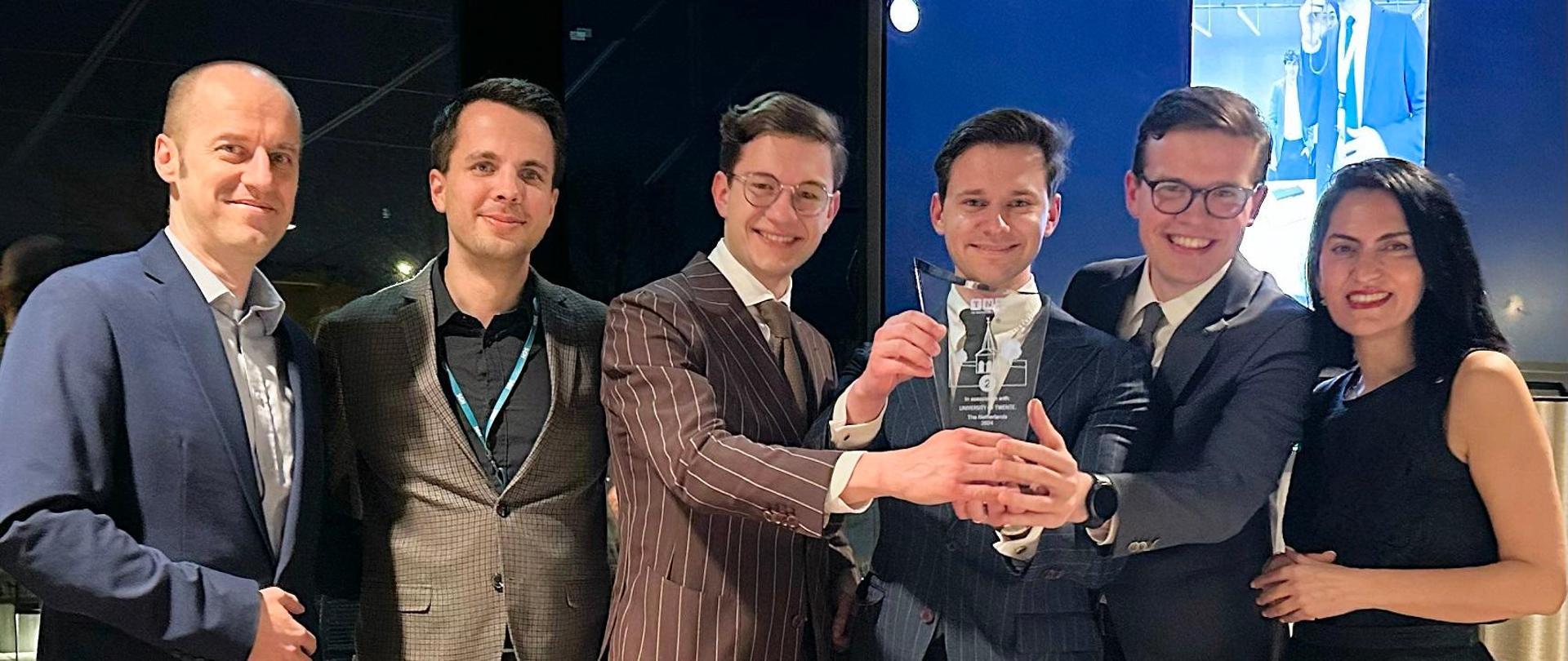 Pięciu mężczyzn w garniturach i kobieta w czarnej sukni stoją w szeregu, trzech pośrodku trzyma szklaną statuetkę.